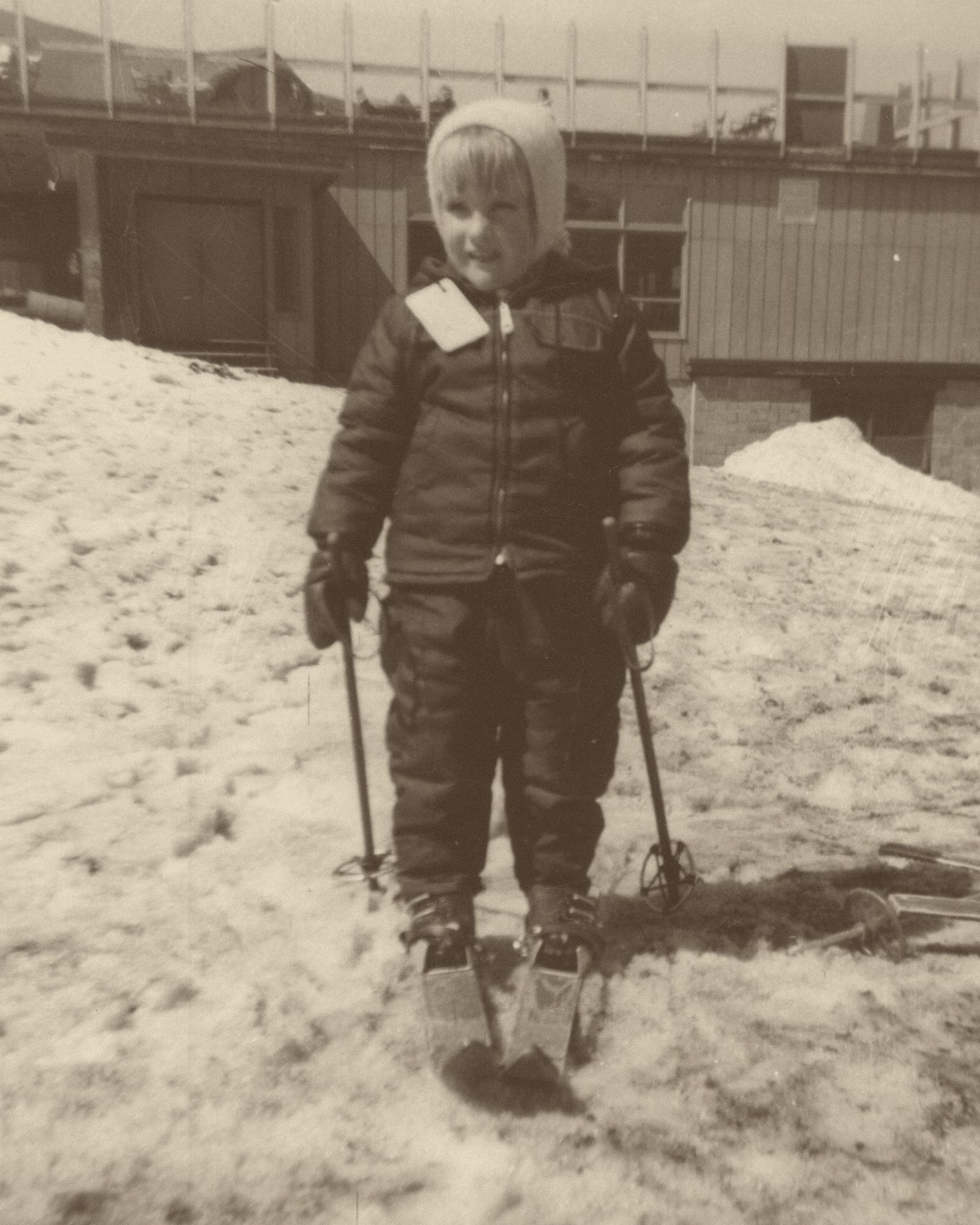 Old sepia photo of Sue as a little girl, standing on skis in a snowsuit and hat in front of a ski lodge